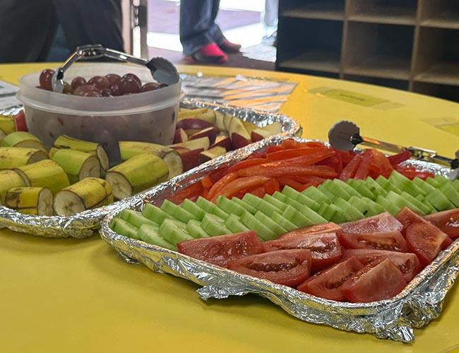 Trays of sliced bananas, apples, grapes, tomatoes, cucumbers, and red bell peppers.